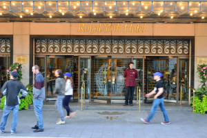 Eine Gruppe von Menschen, die vor dem Royal York Hotel in New York City gehen, mit Text an der Wand, Glastüren, Lichtern und von blühenden Pflanzen umgeben.