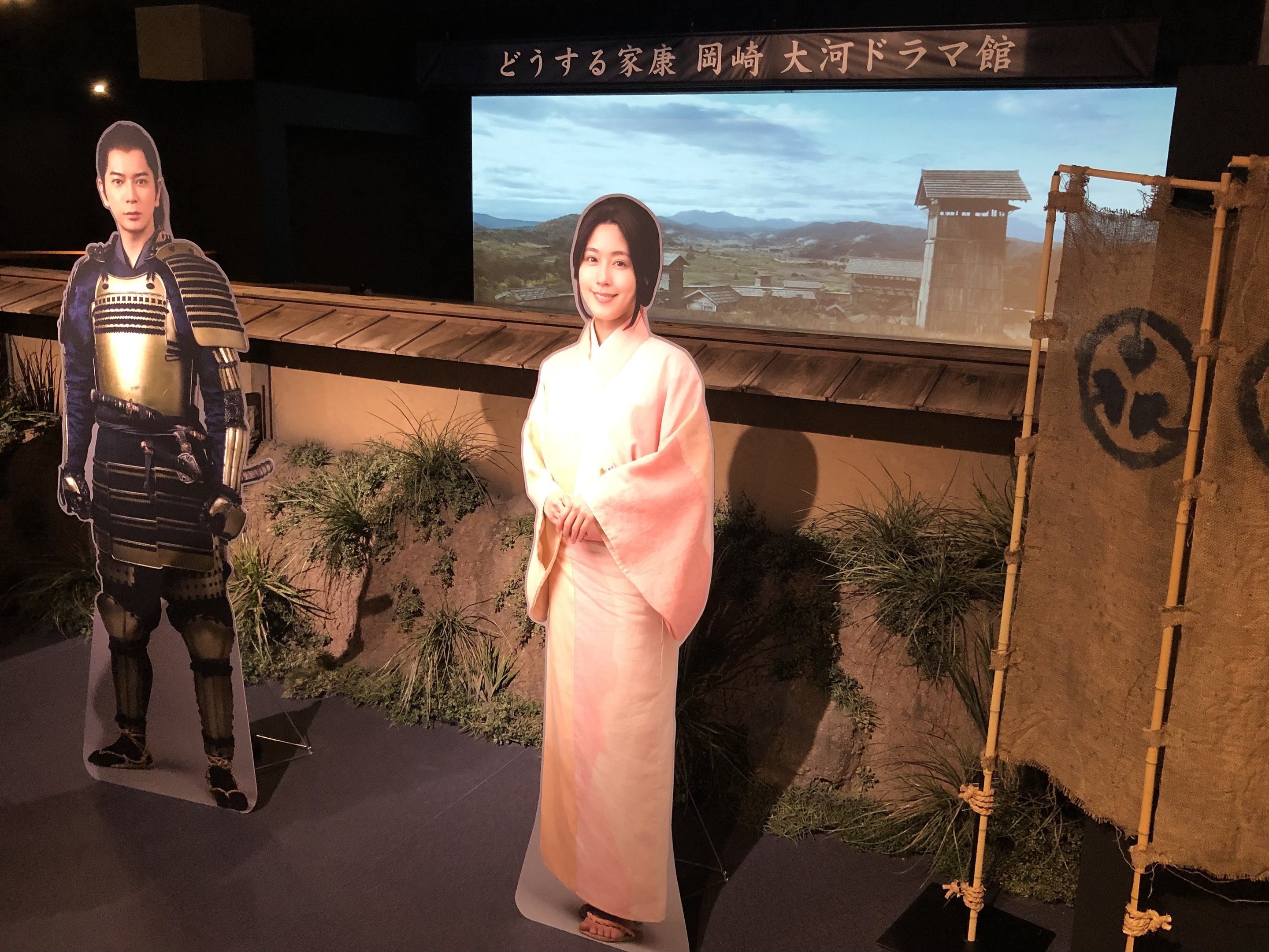 Eine Frau in einem rosafarbenen Kimono und ein Mann in einem Samurai-Kostüm, die zusammen lächeln, auf einem japanischen Filmset mit Pflanzen, einem Bildschirm und Lampen im Hintergrund.