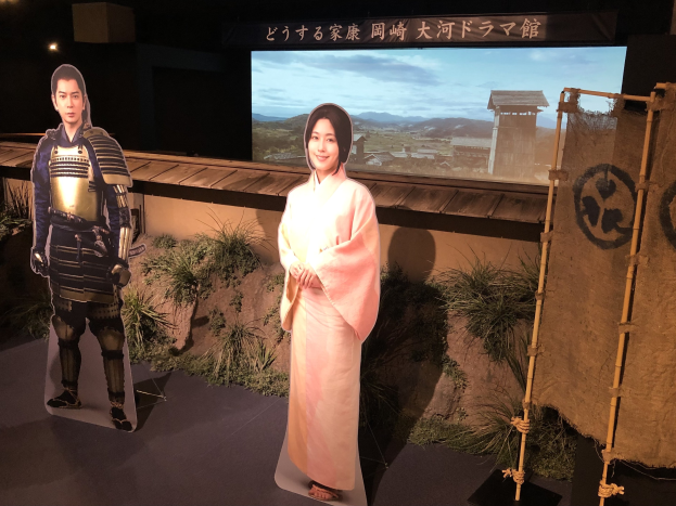 Eine Frau in einem rosafarbenen Kimono und ein Mann in einem Samurai-Kostüm, die zusammen lächeln, auf einem japanischen Filmset mit Pflanzen, einem Bildschirm und Lampen im Hintergrund.