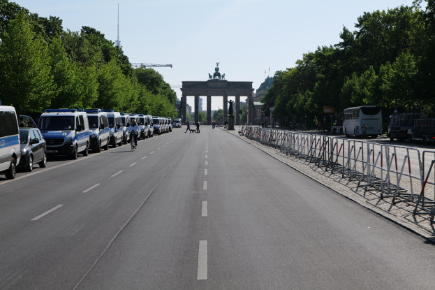 Lange Reihe von Polizeiwagen, die vor dem Brandenburger Tor auf der Straße geparkt sind, mit Fahrradfahrern und Fürdernden, Barrieren, Bäumen und dem Torbogen mit Statuen im Hintergrund.