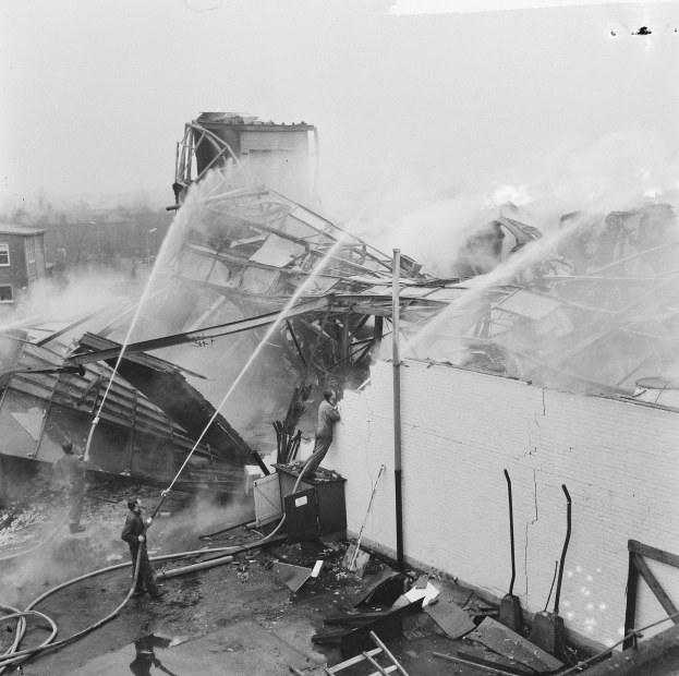 Schwarzes und weißes Foto von Feuerwehrleuten auf einem Dach, die gegen ein Feuer kämpfen, während Menschen unten mit Ausrüstung helfen.