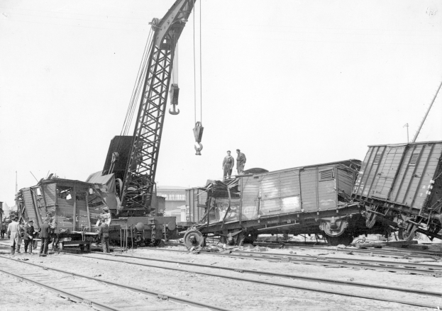 Schwarz-weißes Bild eines Zugunglücks mit einem Kran, der einen Waggon auf die Schienen hebt, mehreren Menschen in der Nähe und Gebäuden im Hintergrund bei klarem Himmel.