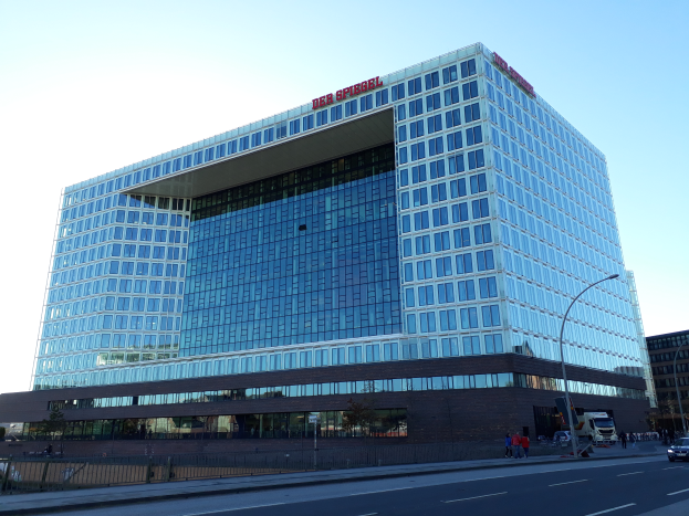 Ein gro├čes Geb├Ąude mit vielen Fenstern, das als Hauptsitz der Deutschen Bank identifiziert wurde, umgeben von Stra├čenmasten, Stra├čenlaternen, Kraftfahrzeugen, Fussg├Ąngern, B├Ąumen und einem bew├Âlktem Himmel.