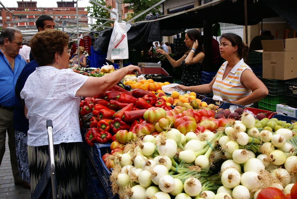 Eine Gruppe von Menschen, die sich draußen in der Nähe von Gebäuden und Bäumen aufhalten, wobei Gemüse in Plastikbehältern und eine Kartonschachtel zu sehen sind.