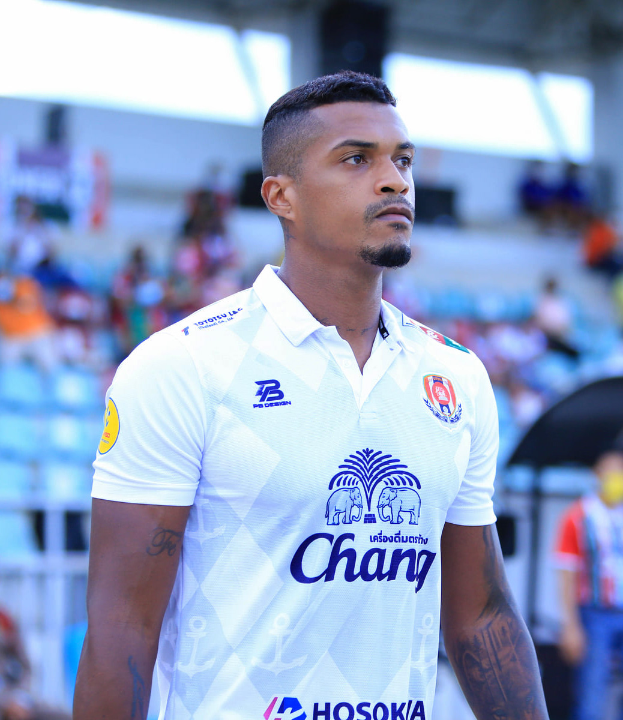 Ein Mann in einem weißen T-Shirt mit Tätowierungen an den Händen steht auf einem Fußballfeld, mit einer unscharfen Gruppe von Menschen im Hintergrund.