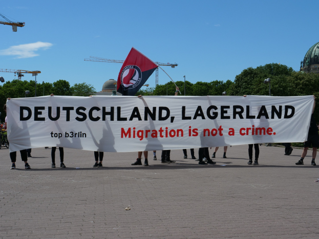 Eine Gruppe von Menschen mit einem Transparent mit der Aufschrift "Deutschland, Lagerland Migration ist kein Verbrechen" und einer Flagge, mit Straßenlaternen, Bäumen, Kränen, einem Gebäude und einem bewölkten Himmel im Hintergrund.