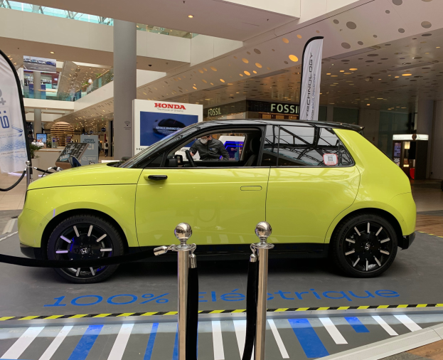 Ein gelbes wasserstoffbetriebenes Elektroauto vor einem Gebäude mit Absperrpollern, Bannern, Menschen, Schildern, einer Topfpflanze, Deckenleuchten und Ventilatoren.