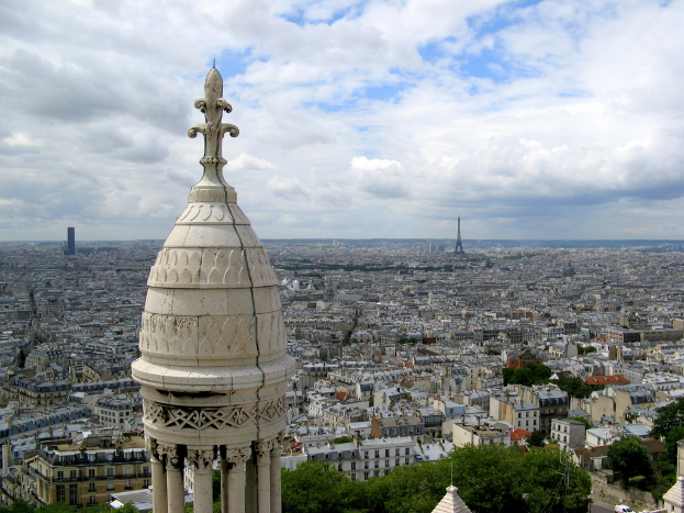 Luftaufnahme einer Stadt mit Gebäuden, Törmen, Bäumen und einem bewölktem Himmel.