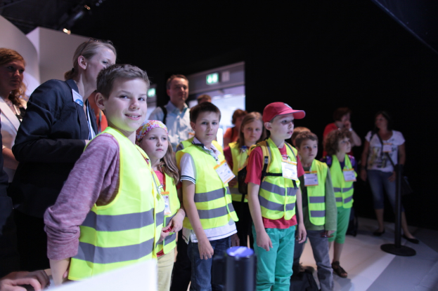 Eine Gruppe von Kindern in Sicherheitswesten und Namensschildern, mit einem Jungen, der links lächelt, auf einem Boden mit einer Stange rechts und ein paar Menschen im Hintergrund.