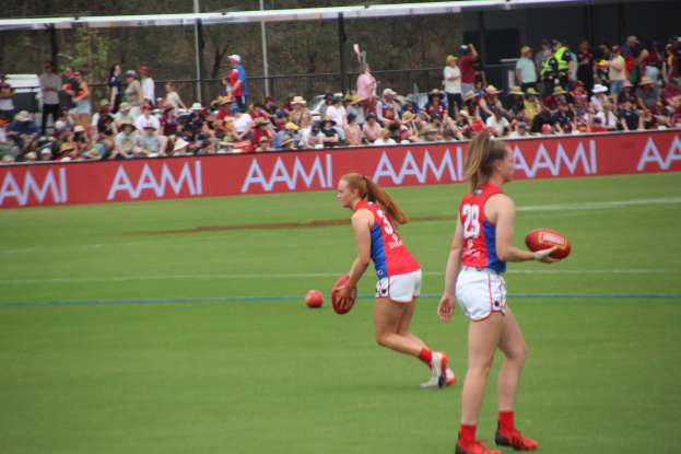 Zwei Frauen in Sportbekleidung spielen Australian Football League auf einem Feld, eine h├Ąlt einen Ball, mit Zuschauern und Tafeln mit Text im Hintergrund.
