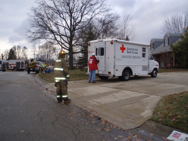 Eine Außenaufnahme mit Bäumen, Rettungswagen, Menschen und einem Gebäude, mit Gras unten und Himmel oben.