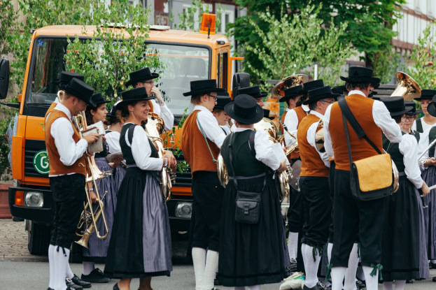 Eine Gruppe von Menschen in traditioneller bayrischer Tracht, die Instrumente spielen und Taschen tragen, mit Bäumen und Gebäuden im Hintergrund und einem geparkten Fahrzeug.