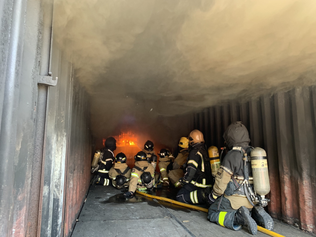 Feuerwehrleute in einem raucherfüllten Tunnel mit Helmen und Sauerstoffflaschen, einer Person mit einer Rohrleitung und einem sichtbaren Feuer im Hintergrund.