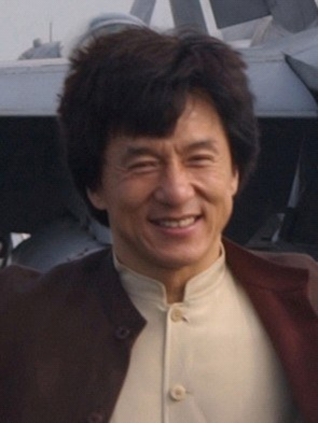 Jackie Chan steht lächelnd vor einem Flugzeug unter einem klaren blauen Himmel, trägt ein weißes Hemd und eine schwarze Jacke mit ordentlich gekämmtem Haar.