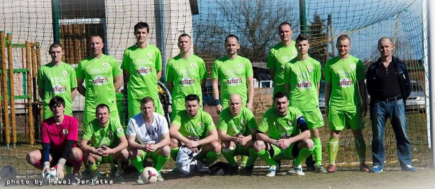 Eine Gruppe von Männern in grünen T-Shirts steht auf einem Fußballfeld in der Nähe eines Balls und eines Torpfostens, mit Bäumen, Gebäuden und Fahrzeugen im Hintergrund.
