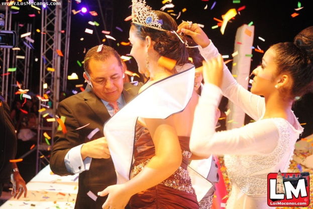 Ein Mädchen auf der Bühne bei einer Miss-World-Show mit einer Krone, dahinter ein Mädchen, das die Krone richtet, und vorne ein Mann in einem schwarzen Anzug, der die Hand schüttelt, vor einem dunklen Hintergrund mit Metallrahmen.