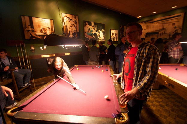 Zwei Personen spielen Snooker an einem von zwei Tischen in einem Club, während mehrere andere im Hintergrund stehen und sich unterhalten, vor einer cremefarbenen Wand mit Fotopostern.