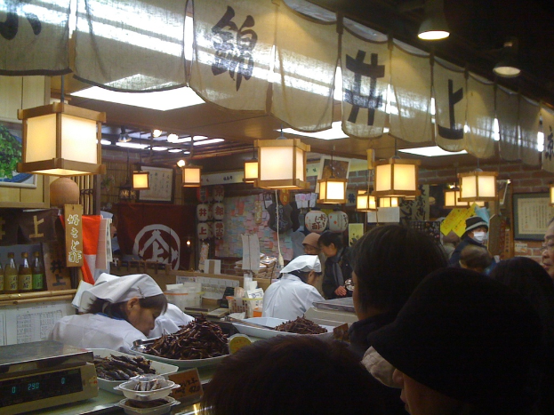 Menschen arbeiten hinter einem Tresen in einem Restaurant mit Gegenständen und Kunden davor und verschiedenen Gegenständen wie Soßenflaschen, Menüs, Kleidung und Lampen im Hintergrund.
