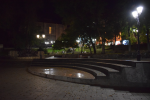 Ein Park bei Nacht mit einem zentralen Brunnen, umgeben von Bäumen und Straßenlaternen, mit einem Gebäude im Hintergrund und Treppen, die zum Brunnen führen.