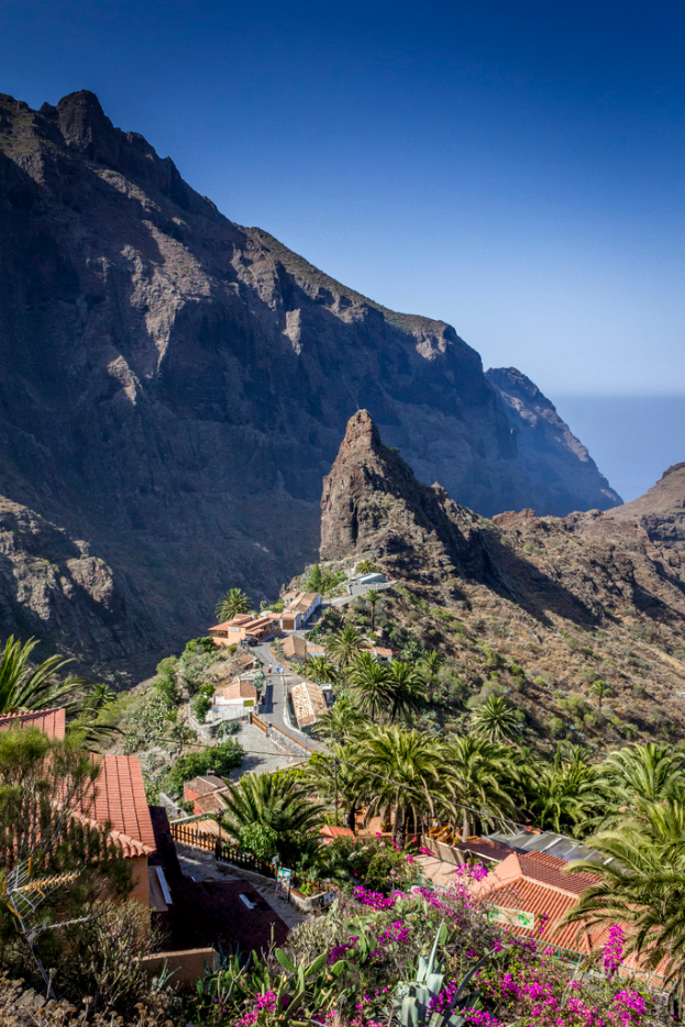 Eine ländliche Szene mit Häusern, Bäumen und Felsen im Vordergrund, einem Hügel im Mittelfeld und dem Himmel im Hintergrund.