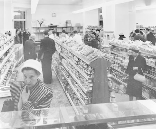 Schwarzes und weißes Foto von Menschen beim Einkaufen in einem Supermarkt mit Lebensmitteln auf Regalen, einige halten Taschen, eine Uhr an der Wand und Deckenleuchten.
