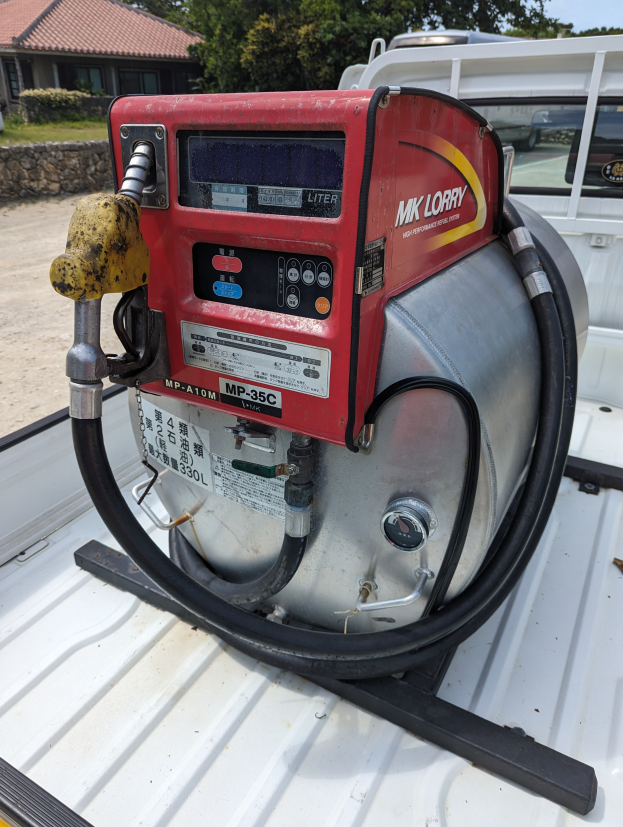 Ein Lastwagen mit einer Tankstelle auf der Ladefläche, umgeben von Bäumen, einem Haus und einem klaren blauen Himmel, ausgestattet mit einem Motor und einem Kraftstoffspender.