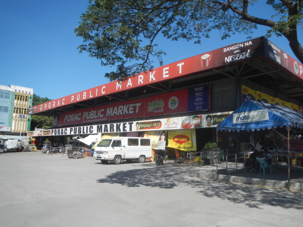 Ein öffentlicher Markt mit einem vor ihm geparkten Van, umgeben von Gebäuden, Geschäften, Schildern, Kraftfahrzeugen auf der Straße, Personen unter einem Zelt sitzend, Geländern, Bäumen und einem klaren blauen Himmel.