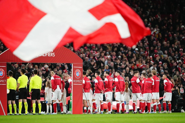 Gruppe von Menschen auf einem Fußballfeld mit einer roten und weißen Flagge im Vordergrund, einem Bogen mit der Aufschrift "Bayern München vs Bayern München Betting Tips & Preview" im Hintergrund und einer großen Menge im Stadion.