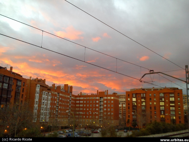 Gebäude mit Fahrzeugen und Bäumen davor, Versorgungsleitungen oben und Himmel im Hintergrund.