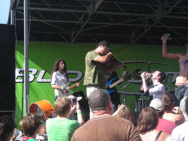 Eine Gruppe von Musikern, die verschiedene Instrumente auf einer Bühne spielen, während das Publikum davor steht und genießt, mit einem großen schwarzen Lautsprecher links und einem grünen Banner im Hintergrund.