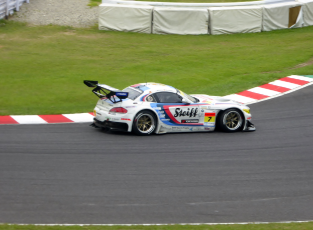 Ein Porsche 911 GT3 RSR fährt auf einer Rennstrecke mit grünem Gras und einem Zaun auf der linken Seite, der mit Text und Zahlen für die 24 Stunden von Le Mans verziert ist.