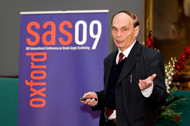 Ein älterer Mann im Anzug hält ein Objekt und spricht, mit einem Banner, einer Wand, einem Foto Rahmen und Blumen im Hintergrund.