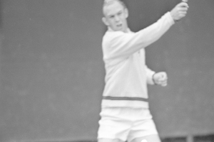 Ein Mann in weißer Tennisbekleidung steht auf einem Tennisplatz und hält einen Schläger in der Hand, mit entschlossener Miene, umgeben von grünem Gras unter einem klaren blauen Himmel.