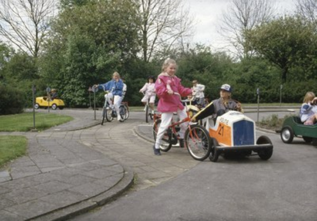 Kinder auf Fahrrädern auf einer von Bäumen gesäumten Straße mit einem Auto und einem Radfahrer im Vordergrund, klarer blauer Himmel im Hintergrund.