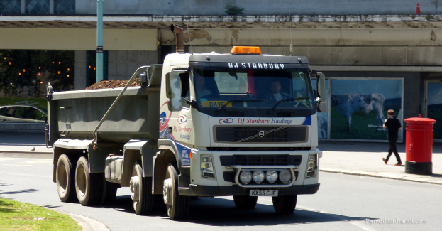 Ein Lastwagen fährt auf einer Straße mit Gebäuden im Hintergrund, einem Briefkasten, einer gehenden Person, einem Pfahl und Grasland.