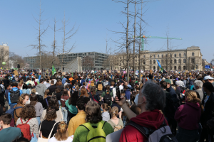 Eine große Menge Menschen mit Schildern und Taschen versammelt sich vor einem Gebäude mit Bäumen und einem klaren Himmel im Hintergrund, was auf eine Klimaprotestveranstaltung hinweist.