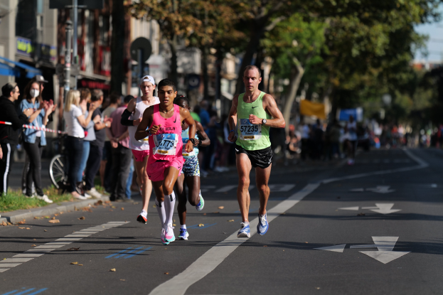 Eine Gruppe von Menschen, die bei einem Marathon auf einer Stadtstraße laufen, mit Zuschauern auf der linken Seite der Straße, Gras am Boden, Bäumen, Gebäuden, Pfosten, Schildern und einem Fahrrad im Hintergrund, alles leicht unscharf.
