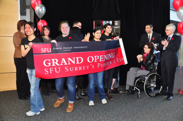 Eine Gruppe von Frauen hält ein Banner hoch, auf dem eine Veranstaltung beworben wird, daneben stehen zwei klatschende Männer und eine Frau im Rollstuhl, die aufgeregt aussieht, vor einem schwarzen Vorhang mit Ballons im Hintergrund.