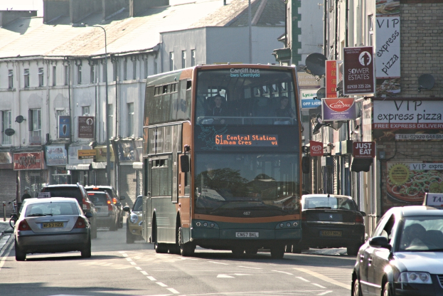 Eine Straße mit Autos und einem Bus vor Gebäuden mit Wänden, Fenstern, Tellern und Dächern, die Plakate und Banner an den Wänden und einen Pfahl mit einer Straßenlaterne zeigen.