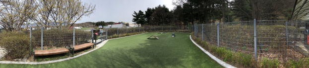 Ein Minigolfplatz in einem Park, umgeben von einem Zaun und grünem Gras, mit Bäumen, Pflanzen, einer Bank, Gebäuden und einem klaren blauen Himmel im Hintergrund.