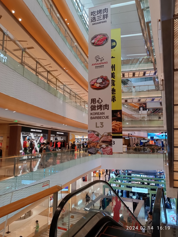 Ein Blick ins Innere eines Einkaufszentrums mit einer Rolltreppe in der Mitte, Menschen mit Taschen unterwegs, Schilder mit Text an der Decke und Geschäften im Hintergrund.