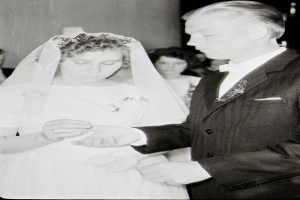 Ein Schwarz-Weiß-Foto einer Braut in einem weißen Kleid und Schleier und eines Bräutigams in einem Anzug, die Ringe austauschen, mit ein paar Menschen im Hintergrund.