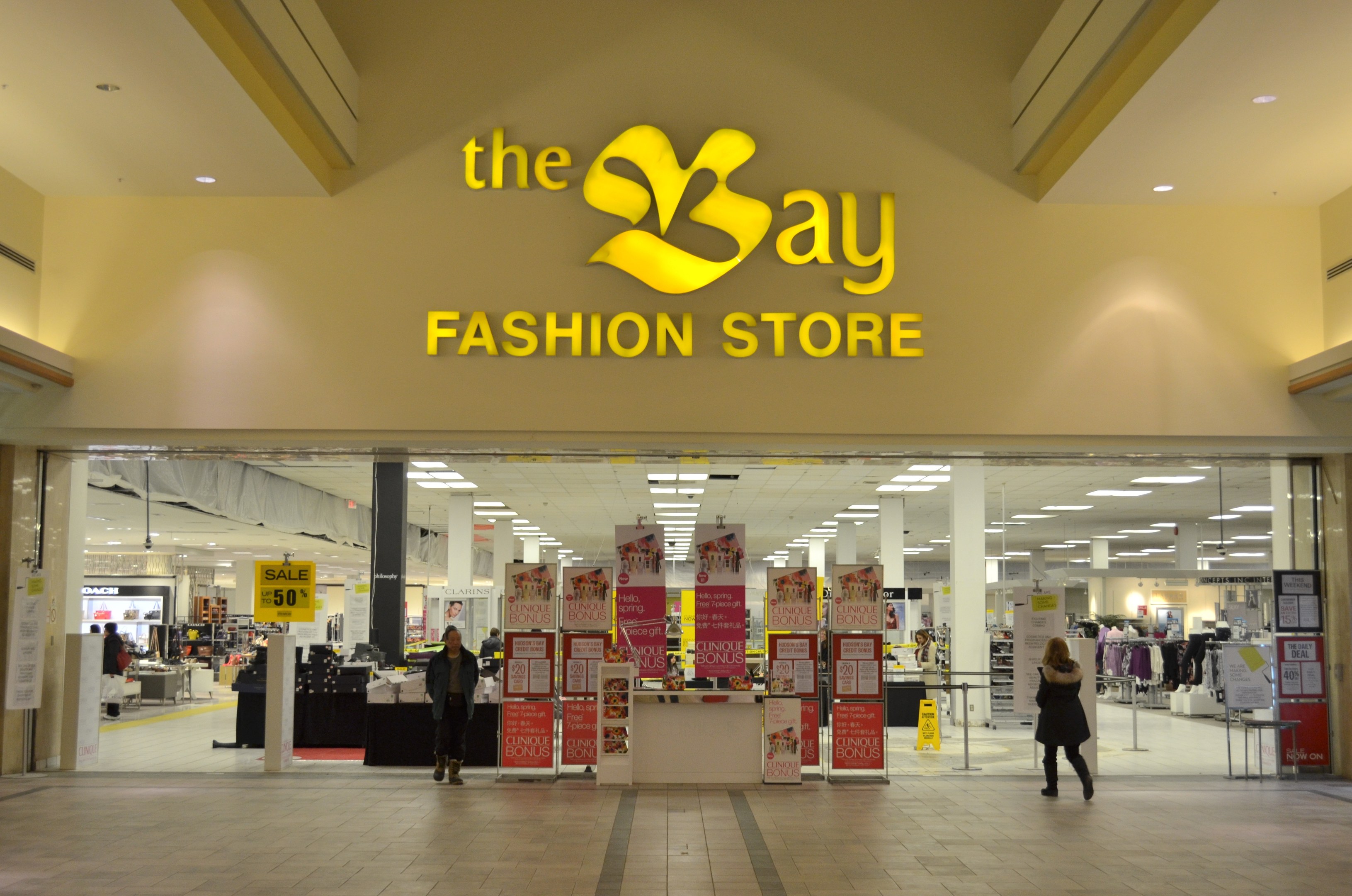 Außenansicht eines Bay Fashion Stores in einem Einkaufszentrum mit Menschen, die gehen, Texttafeln an den Wänden, Säulen, Deckenleuchten und Hintergrundauslagen.
