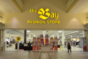 Außenansicht eines Bay Fashion Stores in einem Einkaufszentrum mit Menschen, die gehen, Texttafeln an den Wänden, Säulen, Deckenleuchten und Hintergrundauslagen.