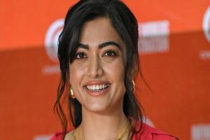 Eine Frau in einem roten Kleid mit einem eleganten Dutt, schwarzen High Heels und natürlichem Make-up lächelt in die Kamera vor einem hellgelben Banner mit der Aufschrift "Sonam Kapoors Beste Schönheits Looks vom Cannes Film Festival."