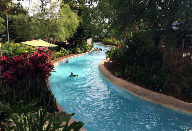 Ein Schwimmbad mit schwimmenden Menschen, umgeben von Pflanzen, Bäumen und einem Zaun, mit Menschen, Schirmen und Gebäuden im Hintergrund.