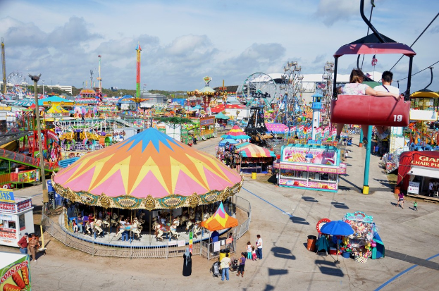Eine Ausstellung mit Fahrgeschäften, Buden, Menschen und verschiedenen Strukturen unter einem bewölkten Himmel.