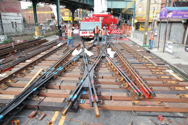 Arbeiter in Jacken und Mützen bauen Schienen, mit einem Fahrzeug, das Material trägt, und einem Schuppen mit Pfählen im Hintergrund und einem Geschäft auf der anderen Seite.