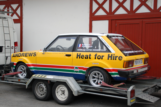 Ein Auto wird auf einem Tieflader von einem Abschleppwagen gezogen, wobei ein weiteres Fahrzeug auf der linken Seite und ein Gebäude mit einer Wand und einer Tür im Hintergrund zu sehen ist.