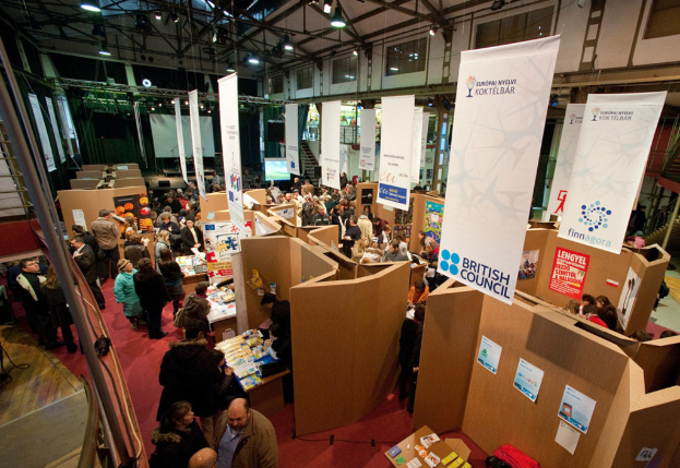 Eine Gruppe von Menschen steht auf einem Teppichboden in einem Raum voller Taesche, Papiere, Gegenstaende, Plakate, Banner, Poster, Bildschirme, Lampen und Glastueren.